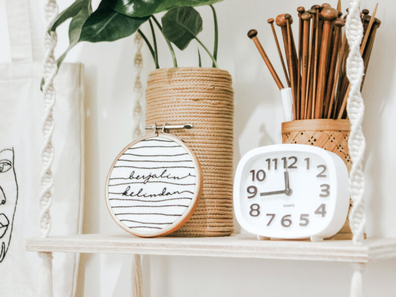 A macrame shelf holds a clock, jar of knitting needles and houseplant in a DIY pot